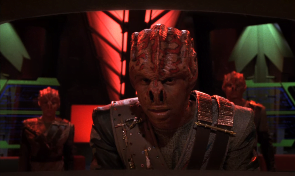 Comandante tamariano em primeiro plano, no telão da Enterprise, com rosto reptiliano e trajes prateados. Atrás dele, no fundo, há outros dois oficiais tamarianos.